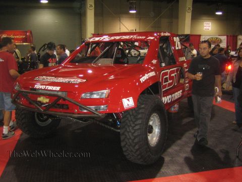 1990 Ford Bronco - red Chevrolet off-road race truck wearing Toyo Tires and number 26 graphics is displayed indoors as people walk past.