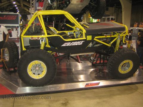 1990 Ford Bronco - Side view of a yellow-and-black tube-chassis rock buggy on large mud tires displayed on an aluminum platform at an expo.