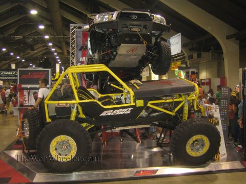 1990 Ford Bronco - yellow-and-black tube-chassis buggy on big mud tires sits on a platform beneath a suspended pickup truck at an indoor show.