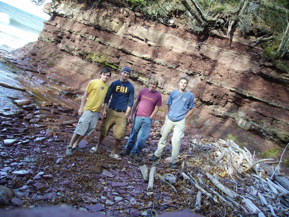 Four guys stand on a rocky shore near a layered cliff face. One wears an FBI shirt