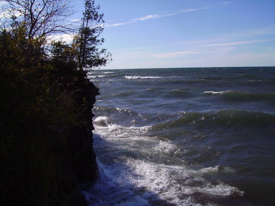 Ocean waves crash against a dark, rocky cliff under a blue sky with wispy clouds