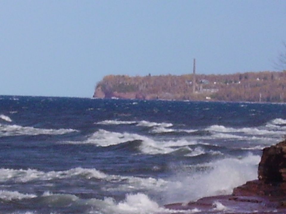 Wavy water with a distant cliffside and smokestack under a clear blue sky