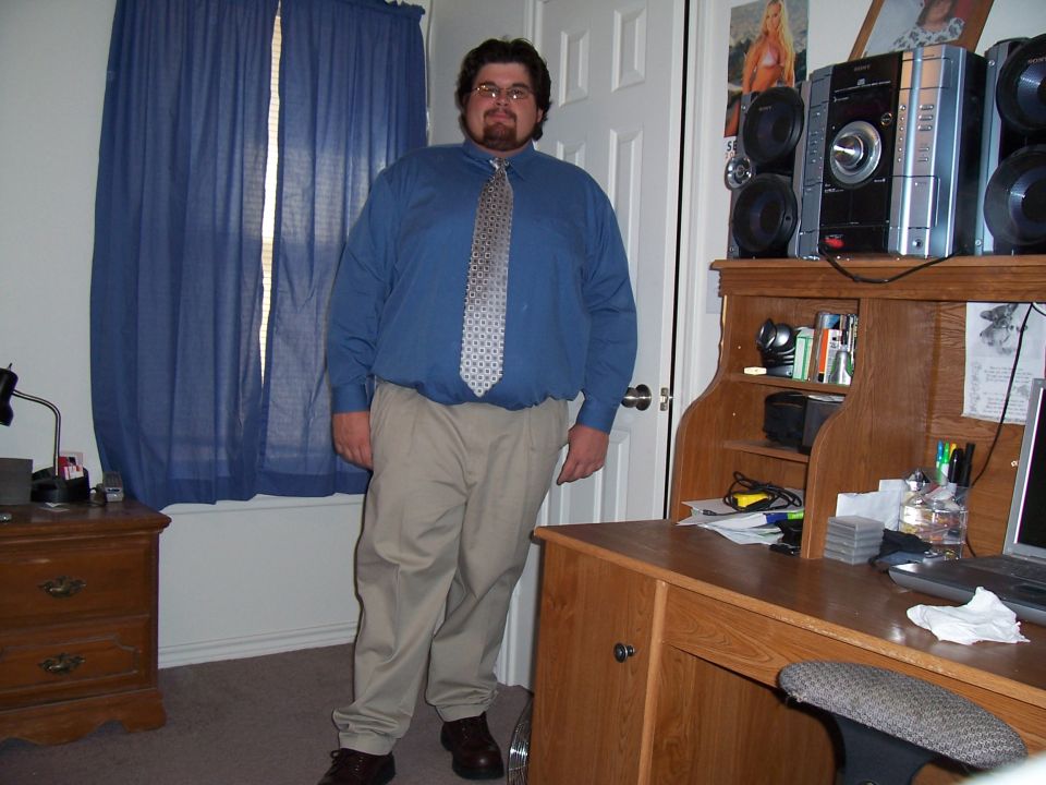 Man in blue shirt, tie, and khaki pants stands in a room with a desk and blue curtains