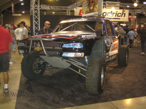 1990 Ford Bronco - Front three-quarter view of a black off-road race truck with tube bumper, BFGoodrich and Lucas graphics on a show floor.