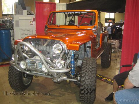 1990 Ford Bronco - orange Larkster Works custom Jeep with a tube front bumper, auxiliary lights and blue coilovers sits at an indoor expo booth.