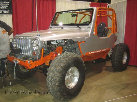 1990 Ford Bronco - silver-and-orange Larkster Juggy custom Jeep on tall mud tires is displayed in a red-curtained expo booth.