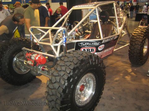 1990 Ford Bronco - bare aluminum tube-chassis rock buggy on big tires, covered in ARB and Turn Key decals, draws onlookers at an indoor show.