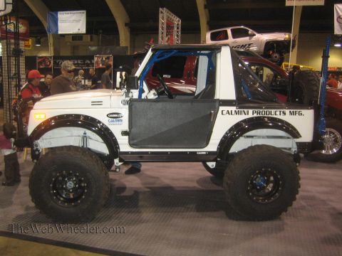 1990 Ford Bronco - Side view of a white Calmini Products lifted 4x4 with a blue roll cage on tall mud tires displayed at an indoor expo.