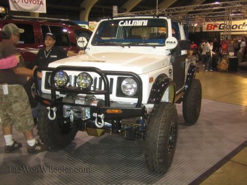 1990 Ford Bronco - white lifted Suzuki-style 4x4 with a winch bumper, auxiliary lights and Calmini graphics sits on a show floor as men inspect it.