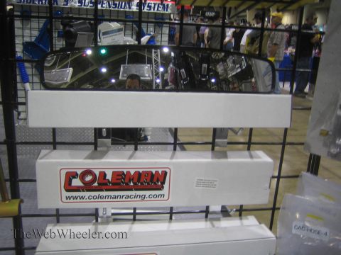 1990 Ford Bronco - wide Coleman Racing rear-view mirror mounted on a diamond-plate stand reflects the show floor at an indoor expo booth.