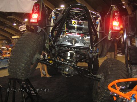 1990 Ford Bronco - black tube-chassis rock buggy raised on stands reveals its rear engine, driveshaft, and solid axle at an indoor event.