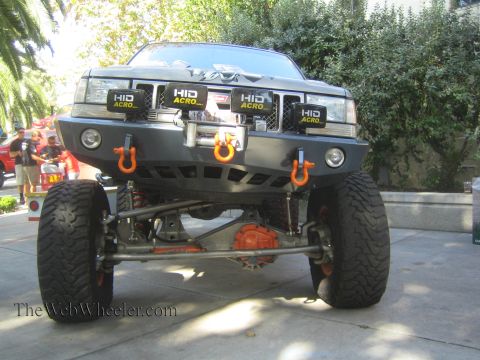 1990 Ford Bronco - lifted gray Jeep Grand Cherokee on big tires shows a custom bumper, HID light pods, and orange tow shackles outdoors.