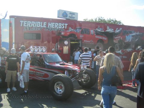 1990 Ford Bronco - red and black Terrible Herbst Motor Sports off-road race car sits before a branded team trailer surrounded by spectators.