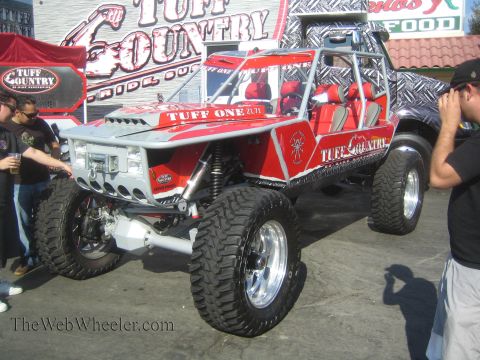 1990 Ford Bronco - red and silver Tuff One off-road buggy on big tires is displayed before Tuff Country suspension banners at an outdoor event.
