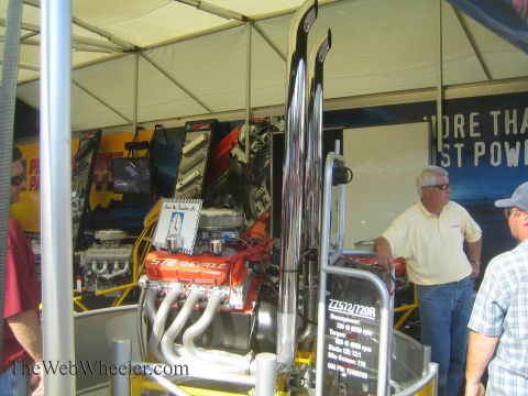 1990 Ford Bronco - red and chrome 572 Chevrolet big-block crate engine is displayed on a stand under a vendor tent as people look on.
