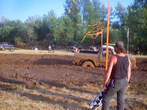 1976 Ford F-100 - Spectator holding a checkered flag watches a truck plow through a mud-bog course outdoors.