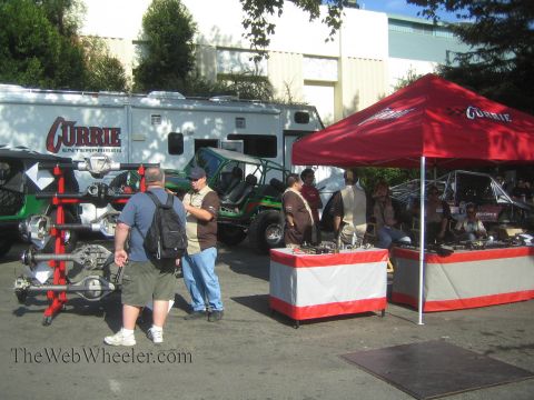 1990 Ford Bronco - Spectators browse a Currie Enterprises outdoor vendor area with a branded trailer, red tents, and an axle display rack.