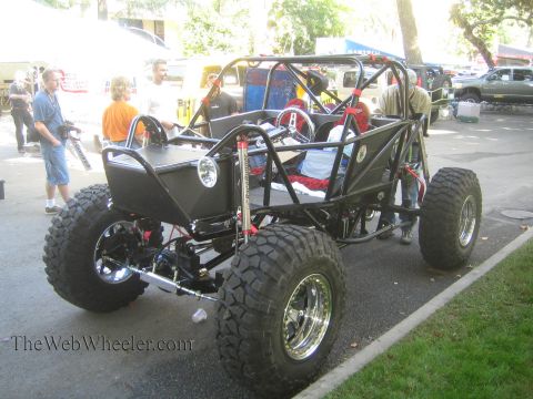 1990 Ford Bronco - black tube-chassis four-seat rock buggy on chrome beadlock wheels is parked on pavement with onlookers nearby.