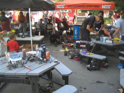 1990 Ford Bronco - Hobbyists work on radio-controlled monster trucks spread across picnic tables in an outdoor pit area under a NOS banner.