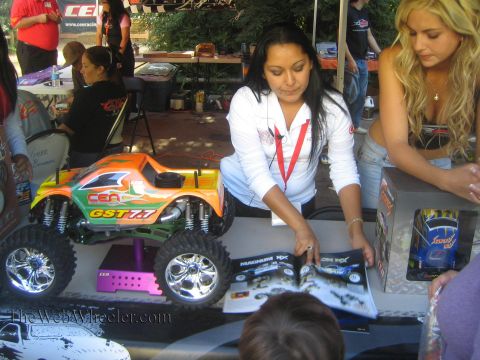 1990 Ford Bronco - woman points to an orange CEN GST 7.7 radio-controlled monster truck displayed beside product boxes at a vendor table.