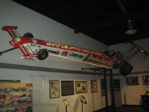 1990 Ford Bronco - red Budweiser King Top Fuel dragster hangs mounted to a wall above framed posters inside the NHRA museum.