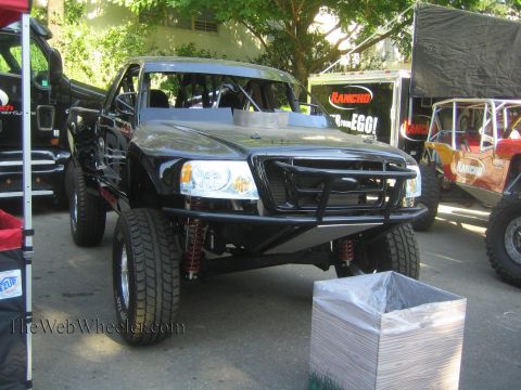 1990 Ford Bronco - black off-road prerunner pickup with a tubular front bumper and roll cage is parked at an outdoor Rancho-sponsored display.