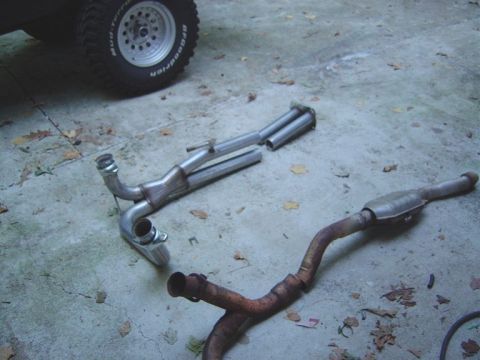 1986 Ford Bronco - Two Y-shaped exhaust assemblies lie on a cracked concrete garage floor: a shiny newer stainless dual-pipe unit with a muffler on top, and a…