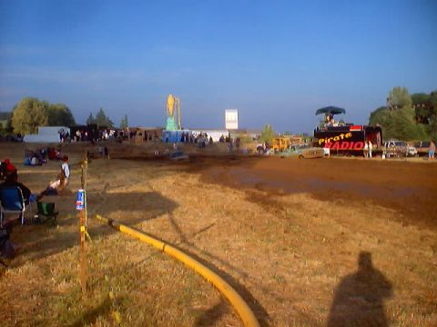 1976 Ford F-100 - An outdoor event is underway with a crowd gathered around a stage and several vehicles visible in the background.
