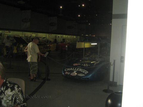 1990 Ford Bronco - dark blue Challenger-lettered dragster sits roped off in a dimly lit museum while a visitor reads a placard nearby.