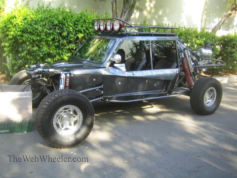 1990 Ford Bronco - black tube-frame off-road buggy with a roof-mounted light bar and For Sale lettering on the body sits beside a hedge.