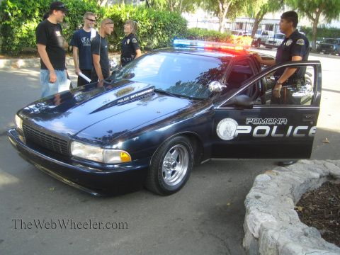 1990 Ford Bronco - dark Pomona Police Chevrolet Caprice cruiser with a roof light bar is parked outdoors as an officer talks with several onlookers.