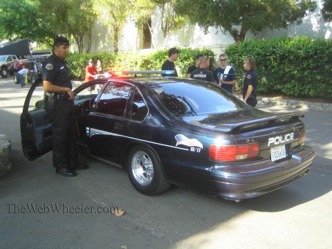 1990 Ford Bronco - Rear three-quarter view of a dark Police-lettered Caprice sedan with a rear spoiler, an officer standing at the open door and people nearby.