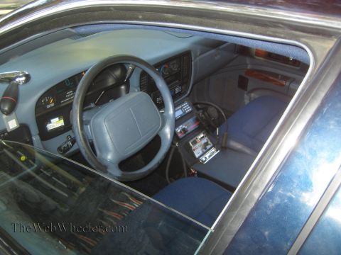 1990 Ford Bronco - Interior view through the window of a police car showing the steering wheel, gauge cluster, blue cloth seats and center-console electronics.