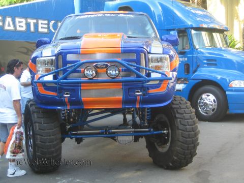 1990 Ford Bronco - Front end of a heavily lifted blue and orange Ford Super Duty truck with a tube bumper and PIAA lights at an outdoor display near a Fabtech…