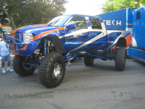 1990 Ford Bronco - Side view of a tall lifted blue Ford crew-cab truck with white and orange graphics on huge tires, parked on pavement at a show.