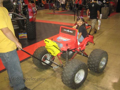 1990 Ford Bronco - young child sits in a red Jeep-style ride-on toy fitted with ATV tires while an adult pulls it across an indoor show floor.