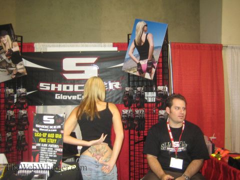1990 Ford Bronco - Shocker GloveCo vendor booth with a branded banner, wall photos and hanging product racks, where a woman speaks with a seated man.