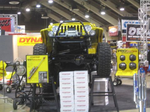 1990 Ford Bronco - yellow tube-fendered off-road Jeep is raised on a display stand at an indoor expo surrounded by Dynatrac product signage.