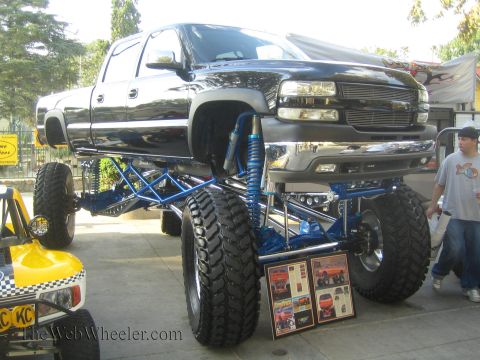 1990 Ford Bronco - black lifted Chevrolet Silverado pickup sits on a tall long-travel suspension with blue coilover shocks at an outdoor display.