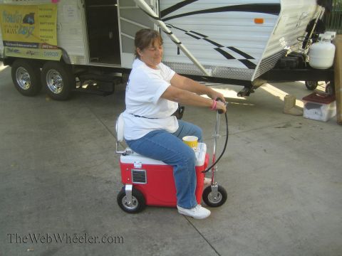 1990 Ford Bronco - woman rides a motorized red cooler-scooter across pavement in front of a parked travel trailer at an outdoor event.
