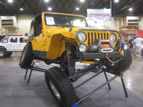 1990 Ford Bronco - yellow Jeep Wrangler rock crawler with a front winch and bumper is raised on a display stand beneath a Toyota banner at an indoor expo.