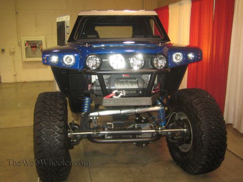 1990 Ford Bronco - Front view of a blue tube-chassis off-road buggy fitted with round light pods and a front winch, displayed indoors against a red curtain.
