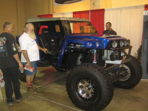 1990 Ford Bronco - blue tube-chassis off-road buggy on beadlock wheels and big tires is displayed at an indoor expo as several men look it over.