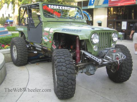 1990 Ford Bronco - green Jeep CJ rock crawler with sponsor decals, red coilover shocks and BFGoodrich tires is parked on pavement outside a venue.
