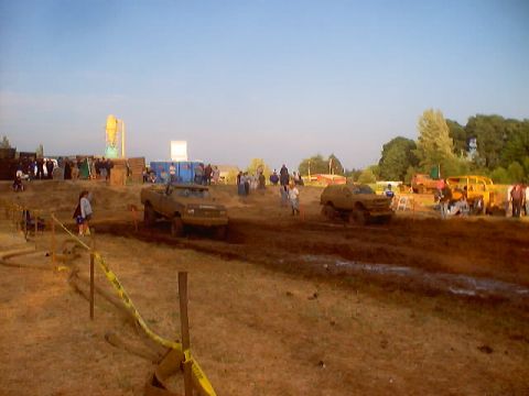 1976 Ford F-100 - Multiple trucks navigate a muddy track during an event, with onlookers gathered in the background.