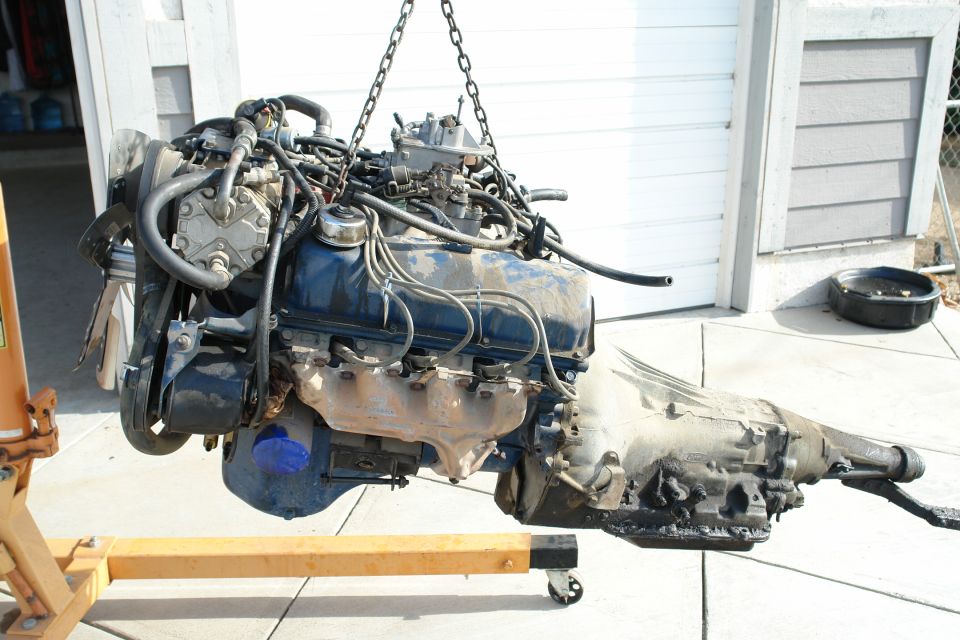 Engine and transmission assembly suspended by chains from an engine hoist, with a black oil pan on the ground