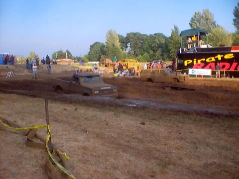 1976 Ford F-100 - A pickup truck navigates a muddy track during a mud run event, with spectators and a trailer visible in the background.