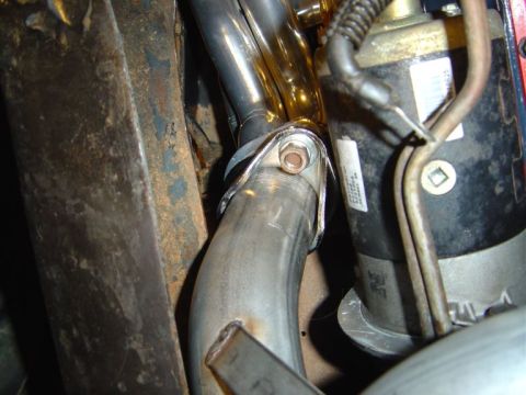 1986 Ford Bronco - Close-up of an exhaust downpipe joint connected with a single visible bolt to a flange, set within a grimy engine bay. Rusted metal bracket…