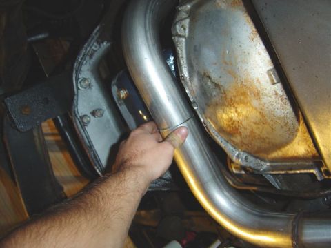 1986 Ford Bronco - Underside view of a stainless steel exhaust pipe routed near a rusted metal shield and black frame bracket, with a hand pointing to the pip…