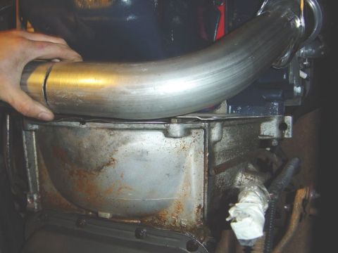 1986 Ford Bronco - Close-up of a hand test-fitting a bent stainless steel exhaust pipe over a rusty metal component near the underside of a vehicle. The pipe…